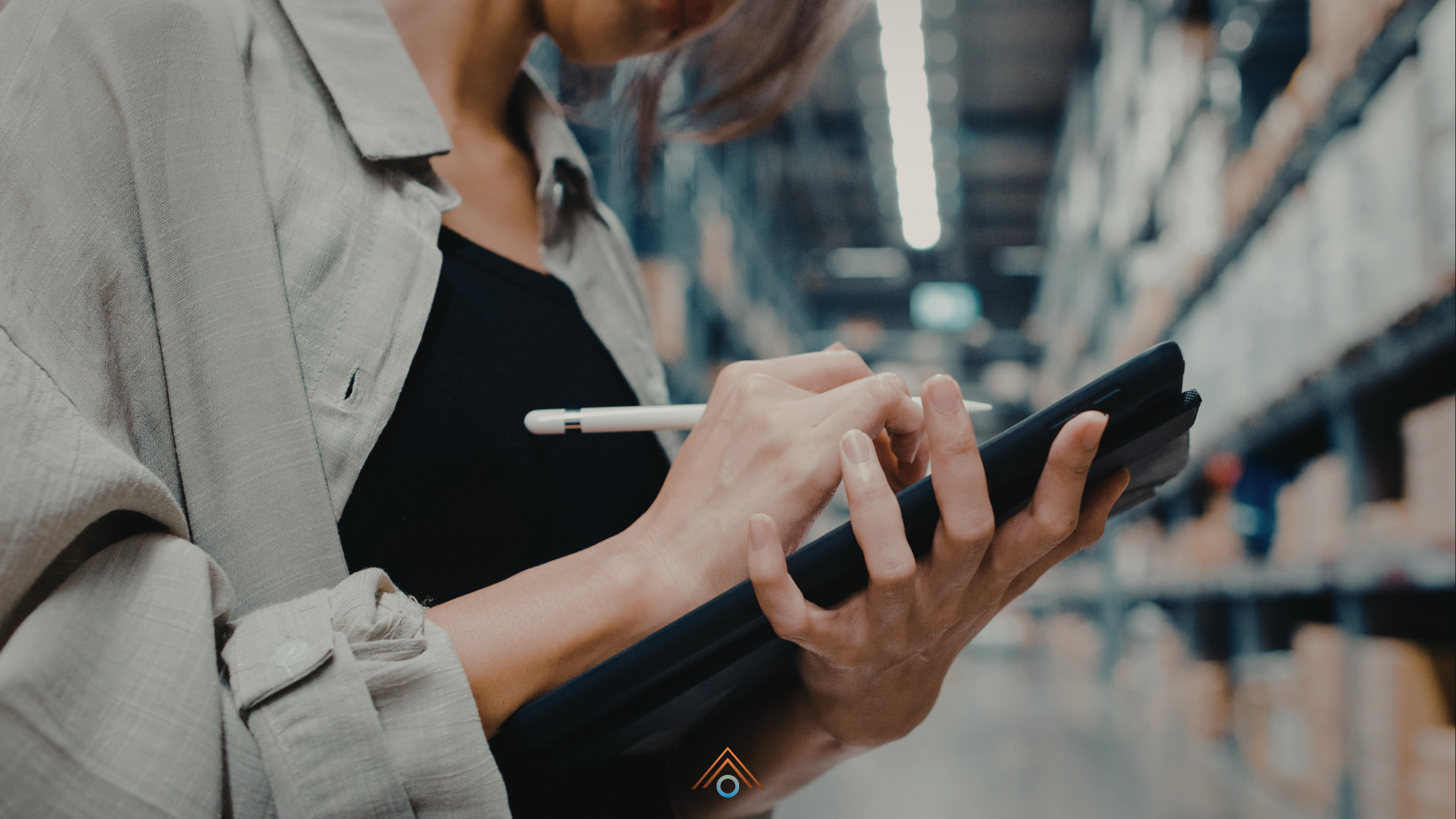 Uma pessoa de close-up, com uma camisa cinza de textura leve sobre uma blusa preta, segura um tablet com capa preta em suas mãos. Ela está usando uma caneta digital branca para fazer anotações na tela do tablet. As mãos e o tablet estão em foco. O fundo é um corredor de armazém de estoque desfocado, com prateleiras altas repletas de caixas de papelão e iluminação fluorescente. Um pequeno logotipo estilizado está no canto inferior central.