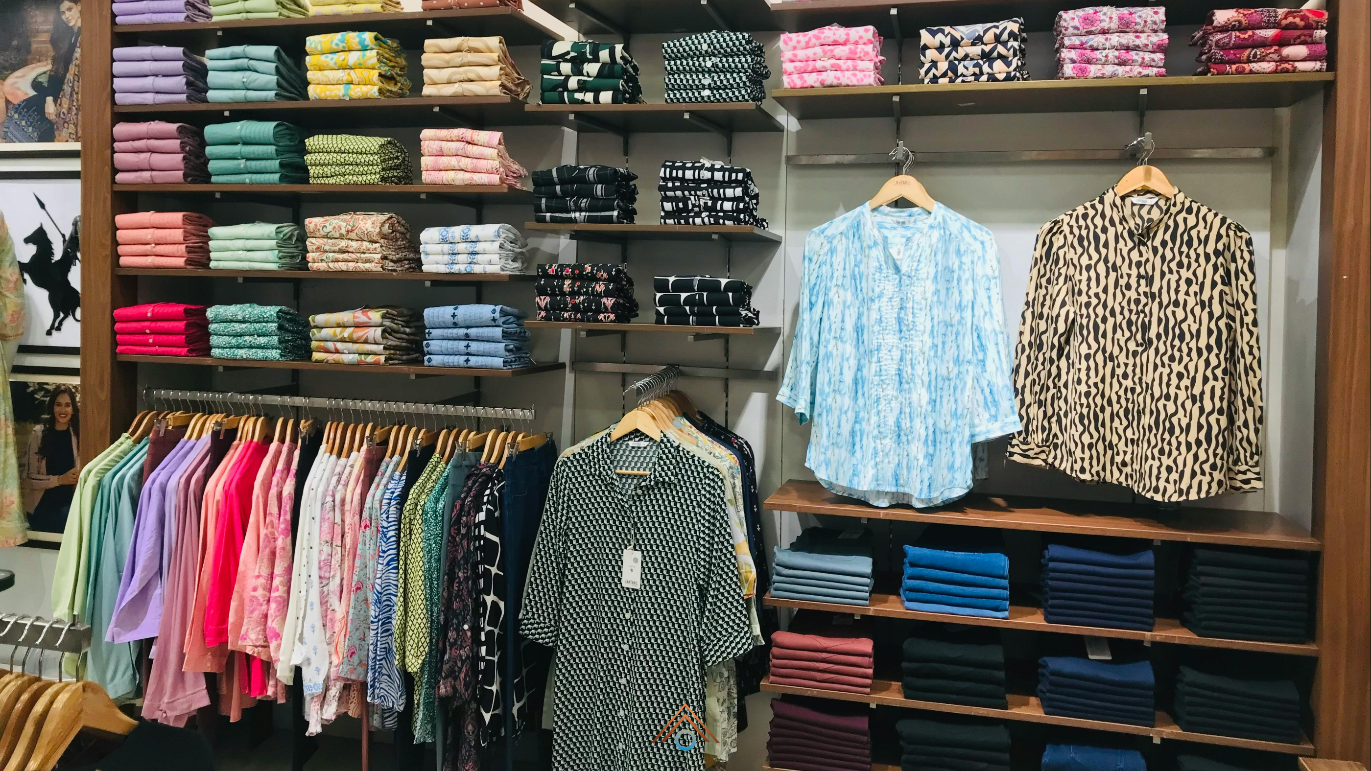 Loja de roupas com prateleiras e araras organizadas, exibindo blusas e camisas femininas dobradas e penduradas em várias cores, estampas e tons pastel, com destaque para duas peças expostas na parede e pilhas de calças jeans dobradas nas prateleiras inferiores.
